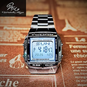 Könnte beinhalten: Eine silberne digitale Casio-Uhr mit einem rechteckigen Zifferblatt, das die Uhrzeit, das Datum und den Tag anzeigt. Die Uhr hat ein Edelstahlarmband und trägt den Text "Casio Multi Lingual Data Bank" und "10 Year Battery". Die Uhr liegt auf einer gemusterten Oberfläche.