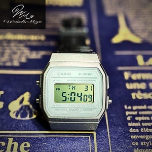 May include: A silver Casio F-91W digital watch with a black band displays the time 5:04:09 PM. The watch face is light green with black text. The watch is on a blue background with white text.