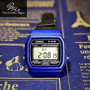 May include: A blue Casio F-91W digital watch with a black strap. The watch face shows the time as 2:08:19. The watch has a rectangular shape and features the Casio logo and model number.
