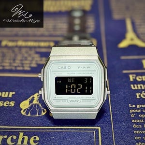 May include: A silver Casio F-91W digital watch with a black digital display showing the time and date. The watch has a light blue face and a silver band. The watch is displayed on a blue surface with gold text.