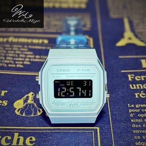 May include: A light blue Casio F-91W digital watch. The watch face displays the time, day, and date. The watch has a light blue band and a rectangular face. The brand name "CASIO" is printed above the digital display.