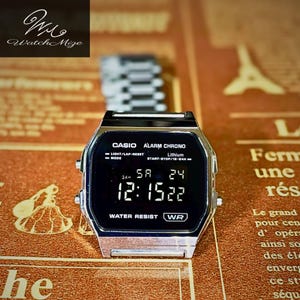 Può includere: Un orologio digitale Casio argentato con un display nero che mostra l'ora, la data e altre funzioni. Il quadrante dell'orologio presenta il logo Casio e le parole "ALARM CHRONO" e "WATER RESIST". L'orologio è su una superficie a motivi.