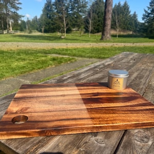 May include: A rectangular wooden cutting board with a hole in the corner, next to a small, round metal tin labeled "PNW PASTE". The board has a rich, warm wood grain pattern. The tin has a silver lid and a yellow label.