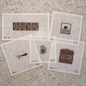 May include: Five pieces of white needlepoint canvas featuring coffee-themed designs. The designs include the text FRESH BREW, JAVA, and COFFEE BEANS, along with illustrations of a portafilter and a French press. Each piece has small color swatches.