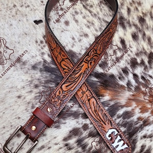 May include: A brown leather belt featuring intricate floral tooling and embossed patterns throughout the strap. The belt includes a dark metal buckle, a matching leather keeper, and the white letters CW embossed on the pointed end of the strap.