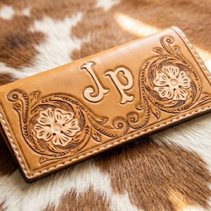 Peut inclure: Un portefeuille en cuir marron clair avec des motifs floraux et des volutes. Le portefeuille porte les initiales "JP" au centre et est posé sur un fond en peau de vache. Le portefeuille est rectangulaire et bordé de coutures.