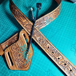 Könnte beinhalten: Ein handgefertigtes braunes Ledergürtel- und Holster-Set. Der Gürtel weist aufwendige florale Prägungen und eine Schnalle auf. Das ebenfalls braune Holster hat passende florale Designs und ist mit gelbem Faden genäht. Das Set wird auf einer grünen Schneidematte präsentiert.