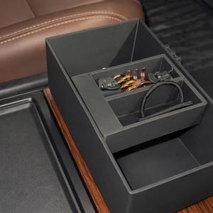 Puede incluir: Un organizador de consola de coche negro con un borde de vetas de madera. El organizador tiene múltiples compartimentos, incluyendo una bandeja con llaves y una correa negra. El interior es negro, y el fondo muestra un asiento de coche marrón.