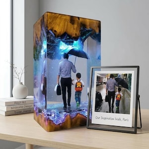Pode incluir: Um bloco de resina retangular com uma cena embutida de uma pessoa e uma criança caminhando sob um guarda-chuva, iluminado com luz azul, ao lado de uma impressão fotográfica com moldura preta com o texto Our Inspiration Walk, Paris.