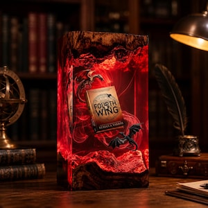 May include: A rectangular resin art piece with a book titled "Fourth Wing" inside. The resin is red with a smoky effect, and the book is flanked by dragon figurines. The piece is set on a wooden surface, with a library background.