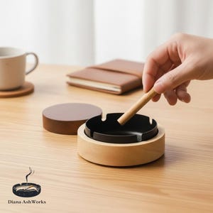 以下が含まれることがあります： 黒い内装と取り外し可能な蓋が付いた木製灰皿。 灰皿は明るい色の木材でできており、円形です。 手が灰皿の上に木の棒を持っています。 ロゴ「Diana Ash Works」が表示されています。