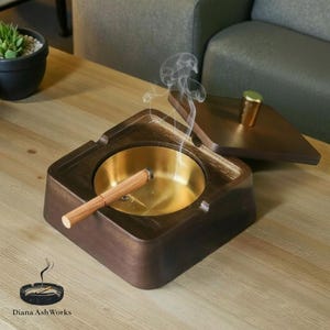 以下が含まれることがあります： 真鍮の内装と蓋が付いた、四角い木製の灰皿。 灰皿には火のついた葉巻があり、煙が立ち上っています。 灰皿は木製のテーブルの上にあります。「Diana Ash Works」のロゴが見えます。