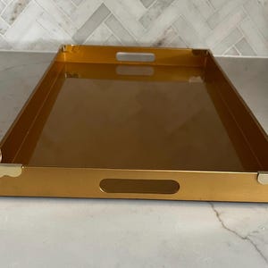 Può includere: Un vassoio rettangolare dorato con maniglie ritagliate sui lati corti. Il vassoio ha una superficie riflettente e angoli dorati. Il vassoio è progettato per servire cibo o bevande.