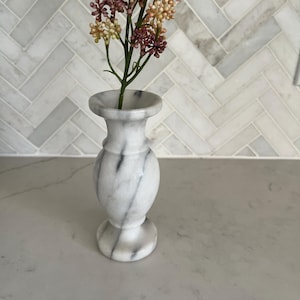 May include: A small, white marble vase with grey veining, featuring a flared rim, a rounded body, and a circular base. A single stem with small, clustered purple and beige flowers is placed inside the opening of the stone vessel.