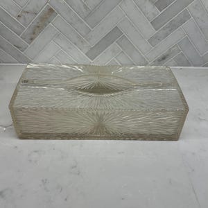 May include: A rectangular clear glass tissue box cover featuring a textured sunburst pattern that radiates from the center of each side and the top. The design includes a central oval opening for dispensing tissues, with a faceted finish.