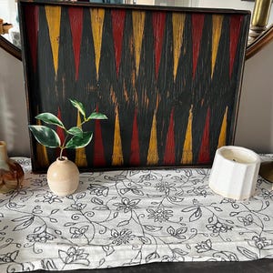 Puede incluir: Una bandeja rectangular de madera con un patrón geométrico negro, rojo y amarillo. La bandeja se exhibe sobre un camino de mesa blanco con estampado floral. También se ven un jarrón pequeño con una planta y una vela blanca.