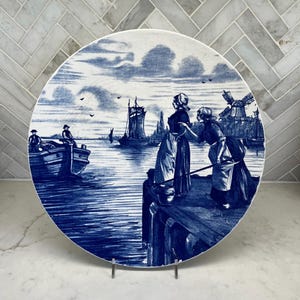 May include: A blue and white decorative plate featuring a nautical scene with figures on a dock, boats, and windmills. The plate has a detailed illustration in a classic Delftware style.