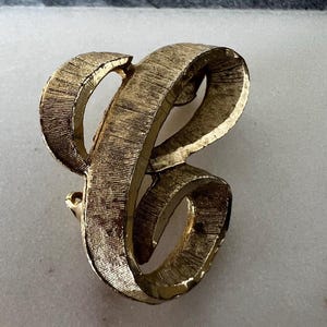 May include: A gold-toned metal brooch in the shape of the letter "B". The brooch has a textured surface and a curved, ribbon-like design. The pin is a vintage accessory.