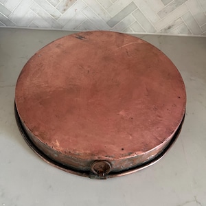 May include: A round copper pan with a flat base and a metallic finish. The pan features a small, circular metal ring handle attached to the side rim. The surface of the copper shows natural variations in color and texture from use and age.