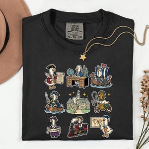 May include: Black Comfort Colors sweatshirt with a gold star necklace and a tan hat. The shirt features colorful cartoon illustrations of ducks in historical and educational settings, including a boat, train, and desk.
