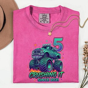 Pu&ograve; includere: Una maglietta rosa Comfort Colors con una grafica di un monster truck verde, il numero 5 e la scritta "Crushing It Since 2021". Una collana d'oro con un ciondolo a stella &egrave; drappeggiata sulla maglietta.