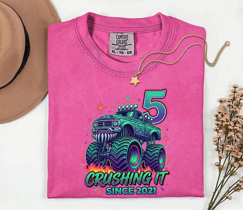 Pu&ograve; includere: Una maglietta rosa Comfort Colors con una grafica di un monster truck verde, il numero 5 e la scritta "Crushing It Since 2021". Una collana d'oro con un ciondolo a stella &egrave; drappeggiata sulla maglietta.