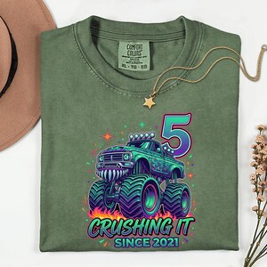 Pu&ograve; includere: T-shirt verde oliva con grafica di monster truck colorato, il numero 5 e la scritta "Crushing It Since 2021". Il design include fiamme, stelle e accenti neon. Visibile anche una collana d'oro con un ciondolo a stella.