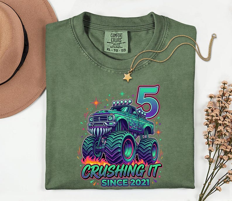 Pu&ograve; includere: T-shirt verde oliva con grafica di monster truck colorato, il numero 5 e la scritta "Crushing It Since 2021". Il design include fiamme, stelle e accenti neon. Visibile anche una collana d'oro con un ciondolo a stella.