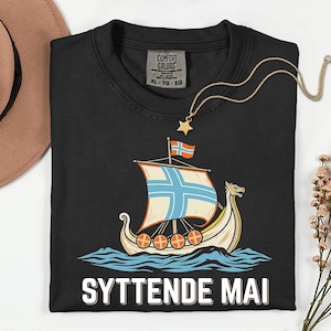 Op de afbeelding: Zwart T-shirt met een afbeelding van een Vikingschip dat op blauwe golven vaart, met de tekst "SYTTENDE MAI" in het wit. Het shirt heeft een Comfort Colors-label en wordt gecombineerd met een gouden ketting.