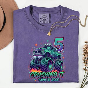Pu&ograve; includere: Maglietta viola con grafica di monster truck verde e turchese, il numero 5 e la scritta "Crushing It Since 2021". Sul davanti una collana d'oro con ciondolo a stella.