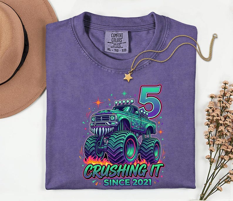Pu&ograve; includere: Maglietta viola con grafica di monster truck verde e turchese, il numero 5 e la scritta "Crushing It Since 2021". Sul davanti una collana d'oro con ciondolo a stella.
