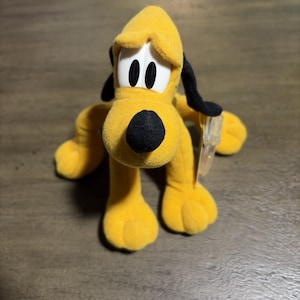 Pode incluir: Um brinquedo de pelúcia amarelo em forma de cão com orelhas e nariz pretos. A personagem tem olhos grandes brancos com pupilas pretas e uma etiqueta de plástico transparente presa na lateral. A pelúcia é de tecido macio e está de pé.