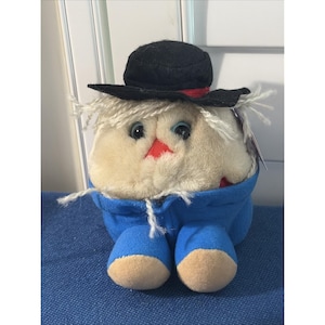Puede incluir: Un juguete de peluche redondo con cuerpo esponjoso de color crema, cabello de hilo blanco, ojos grandes y oscuros y una pequeña nariz roja. El juguete lleva un sombrero de fieltro negro con una cinta roja fina y pantalones de tela azul brillante con pies color canela.