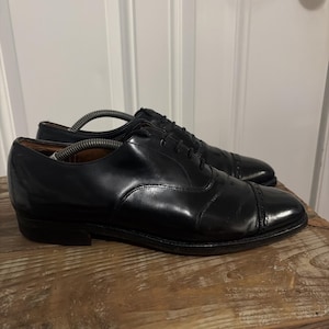 May include: A pair of black leather oxford shoes featuring a cap toe design with subtle decorative perforations along the seam. The shoes have black laces, a low heel, and include internal plastic shoe trees to maintain their structured shape.