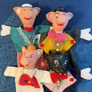 Puede incluir: Un conjunto de cuatro marionetas de mano que representan tres cerdos y un lobo. Los cerdos tienen caras rosadas y diversos atuendos, incluyendo un vestido verde azulado con encaje, un chaleco amarillo con pajarita rosa y una camisa blanca.