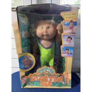 May include: A Cabbage Patch Kids Splash 'n Tan Kids doll in its original packaging. The doll has light brown hair and wears a lime green and purple swimsuit. The box features the text I love the water, BEACH, BATH, POOL, and SPLASH 'N TAN KIDS.