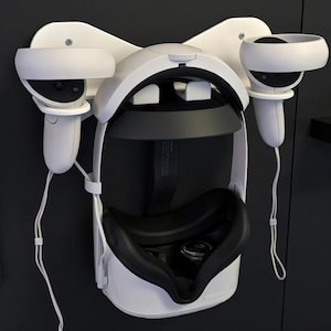 May include: White VR headset and controllers mounted on a wall-mounted holder. The headset has a black interior and is flanked by two controllers. The holder is white and designed to cradle the headset and controllers.
