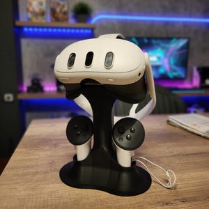 Op de afbeelding: Een witte en zwarte VR-headset met drie naar voren gerichte lenzen, rustend op een zwarte standaard met twee aangesloten controllers. De standaard staat op een houten oppervlak. De headset heeft een strak, modern ontwerp, wat duidt op virtual reality gaming of entertainment.