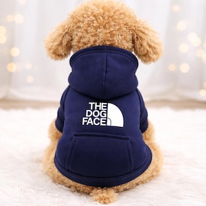May include: A navy blue dog hoodie with a white graphic that reads "THE DOG FACE". The hoodie has a hood and a front pocket. The dog is a light brown color.