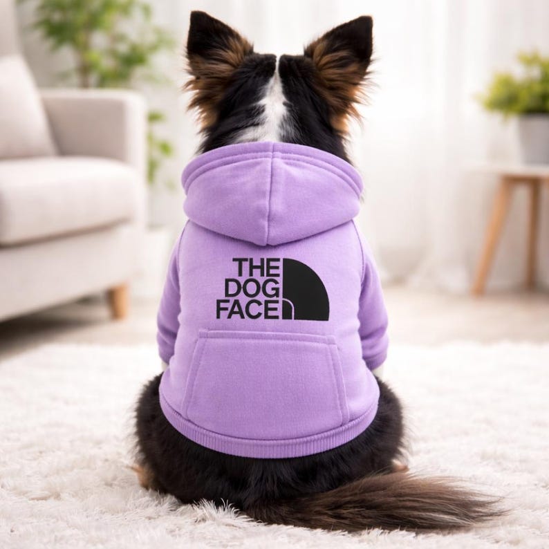May include: A lilac-colored dog hoodie with a black graphic that reads "THE DOG FACE". The hoodie has a kangaroo pocket and a hood. The dog is sitting on a white rug.