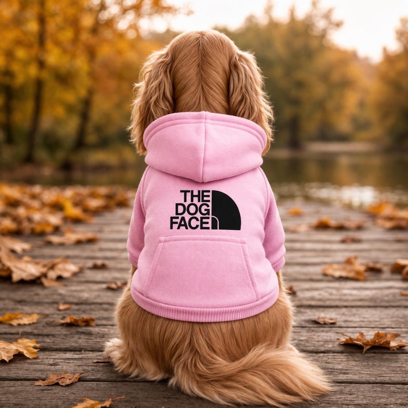 May include: A light pink dog hoodie featuring a hood, long sleeves, and a kangaroo pocket. The back of the garment displays the black text THE DOG FACE alongside a black semi-circular graphic logo printed on the center of the fabric.