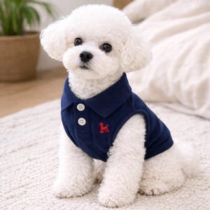May include: A small, white dog wearing a navy blue polo shirt with a red embroidered emblem. The shirt has a collar and two white buttons. The dog is sitting on a white, textured surface.
