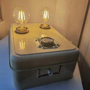 Puede incluir: Una caja de metal color crema con dos bombillas de estilo Edison en la parte superior. Las bombillas emiten un brillo amarillo cálido. La caja tiene un pequeño indicador y un botón plateado. La caja mide unos 30 cm de largo y está sobre una superficie blanca.