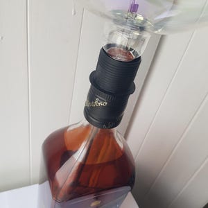 May include: A repurposed lamp made from a Johnnie Walker Black Label whiskey bottle. The bottle is filled with amber liquid and topped with a black lamp socket and a large, clear light bulb. The lamp is a unique piece of home decor.