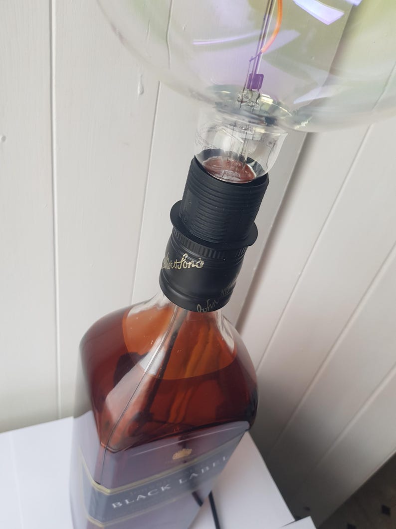 May include: A repurposed lamp made from a Johnnie Walker Black Label whiskey bottle. The bottle is filled with amber liquid and topped with a black lamp socket and a large, clear light bulb. The lamp is a unique piece of home decor.