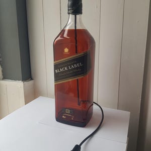 May include: A repurposed Johnnie Walker Black Label whiskey bottle transformed into a lamp. The amber-colored glass bottle features a black label with gold lettering. A black lamp socket and cord with an on/off switch are attached.