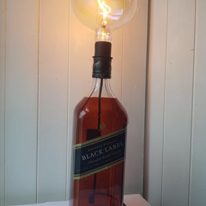 May include: A unique lamp made from a repurposed Johnnie Walker Black Label bottle. The brown glass bottle is topped with a black lamp holder and a large, illuminated Edison-style light bulb. The lamp is plugged into a wall outlet.
