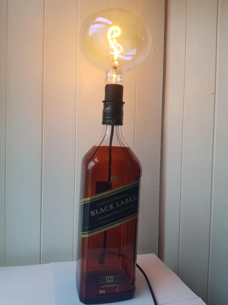 May include: A unique lamp made from a repurposed Johnnie Walker Black Label bottle. The brown glass bottle is topped with a black lamp holder and a large, illuminated Edison-style light bulb. The lamp is plugged into a wall outlet.