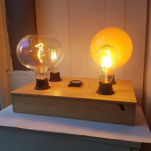 May include: A wooden box with three decorative light bulbs. Two large, clear bulbs and one smaller, teardrop-shaped bulb are lit, casting a warm glow. The bulbs are mounted on black bases. A yellow globe is behind the teardrop bulb.