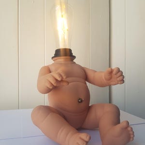 May include: A table lamp featuring a seated baby doll figure with a light bulb replacing the head. The doll has a light peach skin tone and a metal toggle switch located on the stomach area. The clear glass light bulb emits a warm glow.